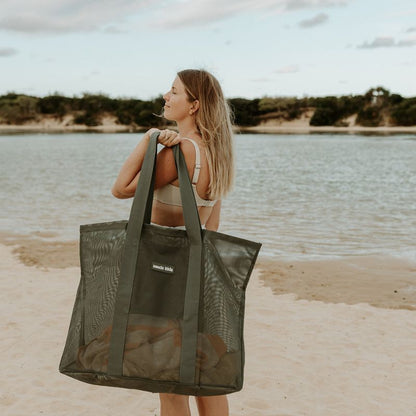 A woman at a lake beach carrying the sande kids oversized mesh beach bag in olive sage green