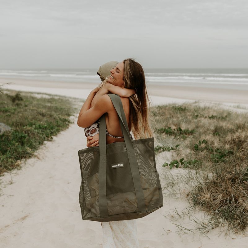 Mother at the beach carrying her toddler son with a Sande Kids Beach Hauler Mini in Pandanus green on her shoulder.