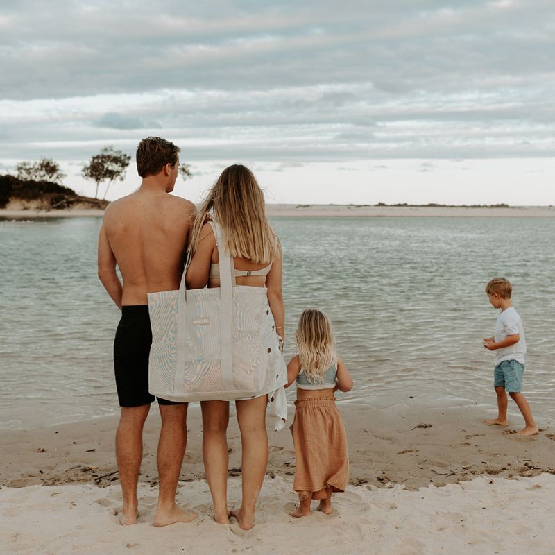 Family of four at the beach. Mum stadning next to Dad while their little boy and girl play. Mum carries the Sande Kids Beach Hauler mesh beach bag in neutral.