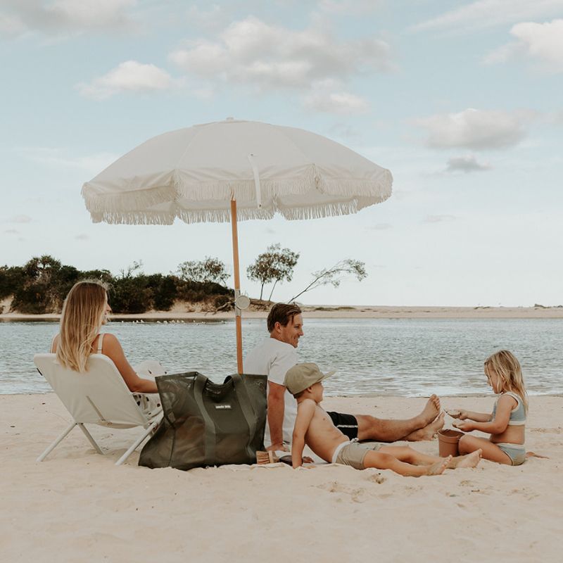 A young family hanging a a lake beach. White Basil Bangs and beach chair, family lazing arounds. A Sande Kids Beach Hauler mesh beacg bag in Pandanus feint and centre.