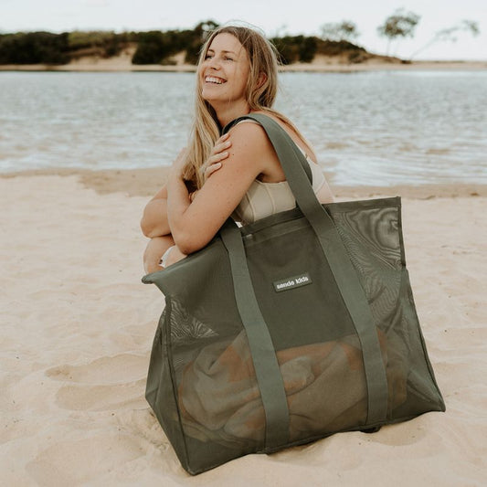 A woman crouched next to the Sande Kids oversized mesh beach bag in green.