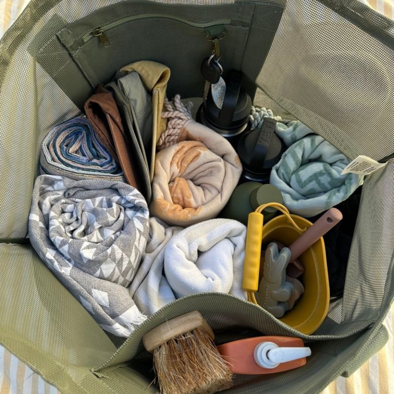 Image to demonstrate what fits in to the Sande Kids Beach Hauler - oversized mesh beach bag. Colour is Pandanus green. Packed inside are beach towels, beach toys, sand brush, sunscreen, water bottles.