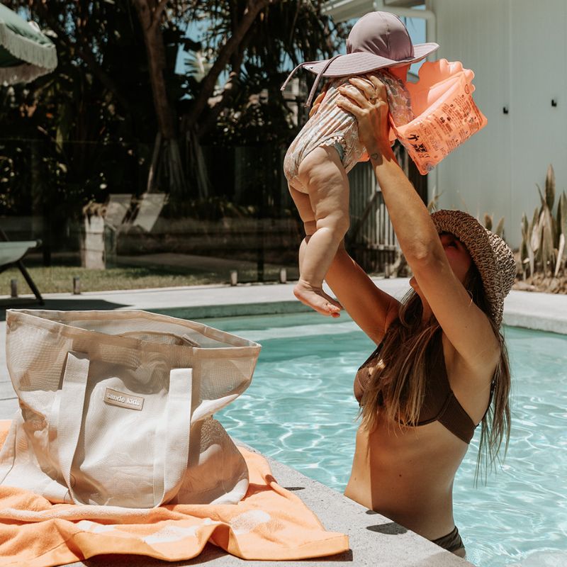 Mum in a beautiful backyard pool with her baby girl. Image features the Sande Kids original mesh beach bag in Sand sitting on a peach coloured beach towel.