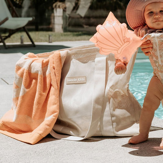Baby girl at the pool with pink shell floaty bands. Image features the Sande Kids Beach Hauler mesh bag in Sand.