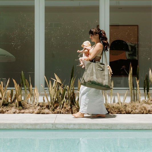 Mother walking past a pool with an olive green mesh beach bag on one shoulder and a newborn baby in her arms.