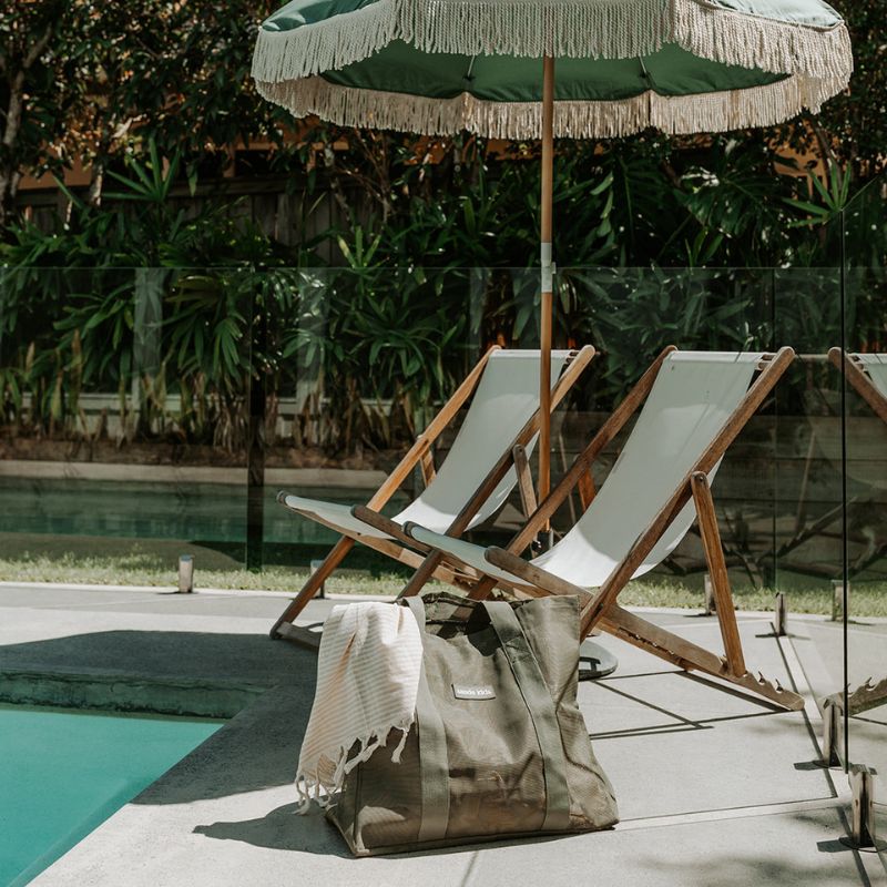 A backyard pool scene, two canvas beach chairs, a green beach umbrella with cream tassels in an umbrella stand and a Sande Kids Beach Hauler Mini in Pandanus green.