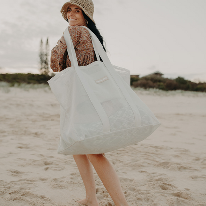Woman at the beach smiliing. Wearing a crochet straw hat and brown fine floral printed dress. Carrying a Sande Kids Beach Hauler family beach bag.