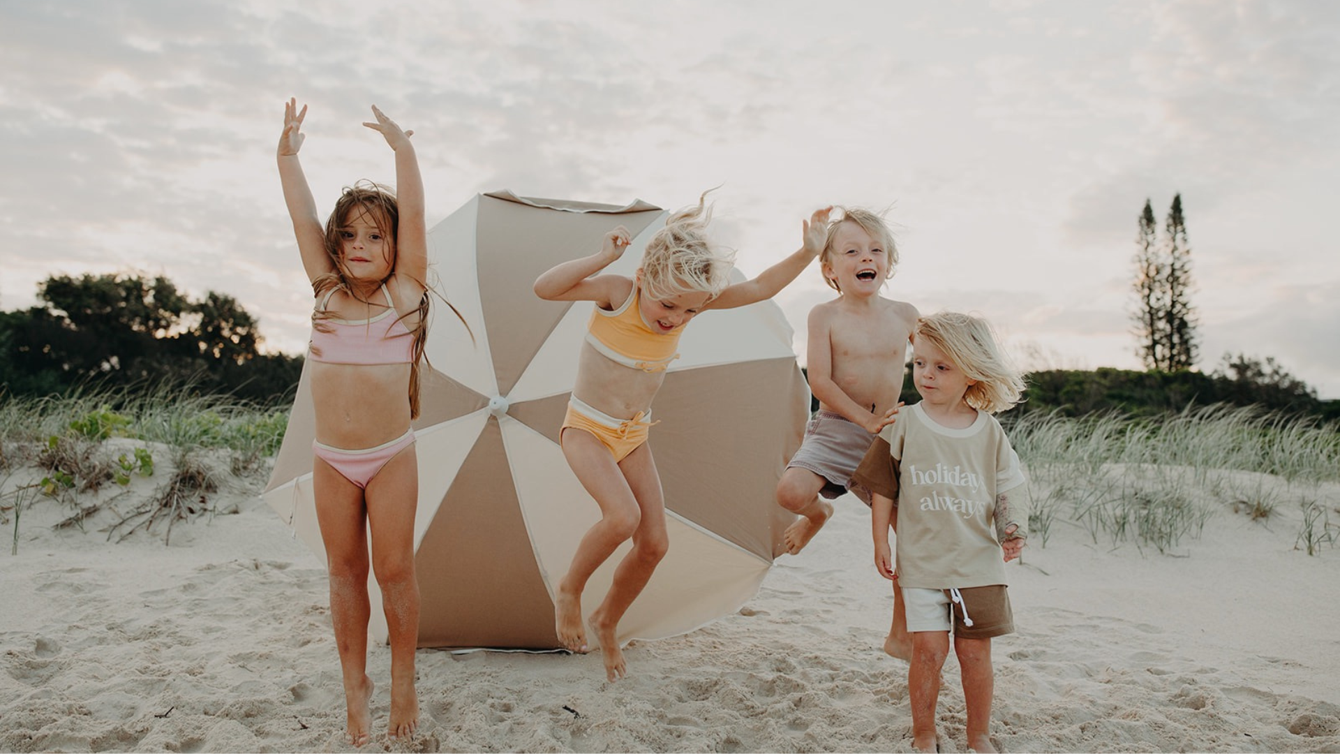 Four children at the beach jumping for joy in front of a beige and cream beach umbrella with coastal dune in the back ground. There is little brunette girl dressed in a pink bikini, a little blonde girl dressed in a yellow bikini, a shaggy blonde haired boy in grey shorts and a very little boy with blonde surfer hair where a brown and tan t-shirt and shorts. The t-shirt says Holidays Always. 